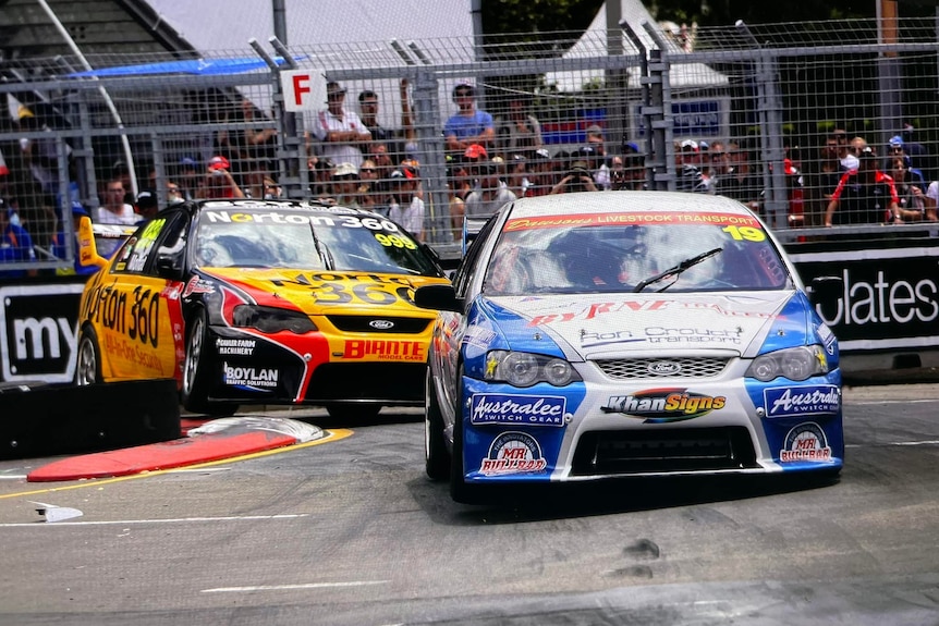 Two V8 supercars plastered with their sponsors' logos round a hairpin bend as a crowd watches on.