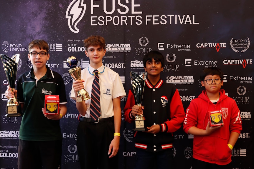 four students posing in front of a sponsorship wall with their trophies