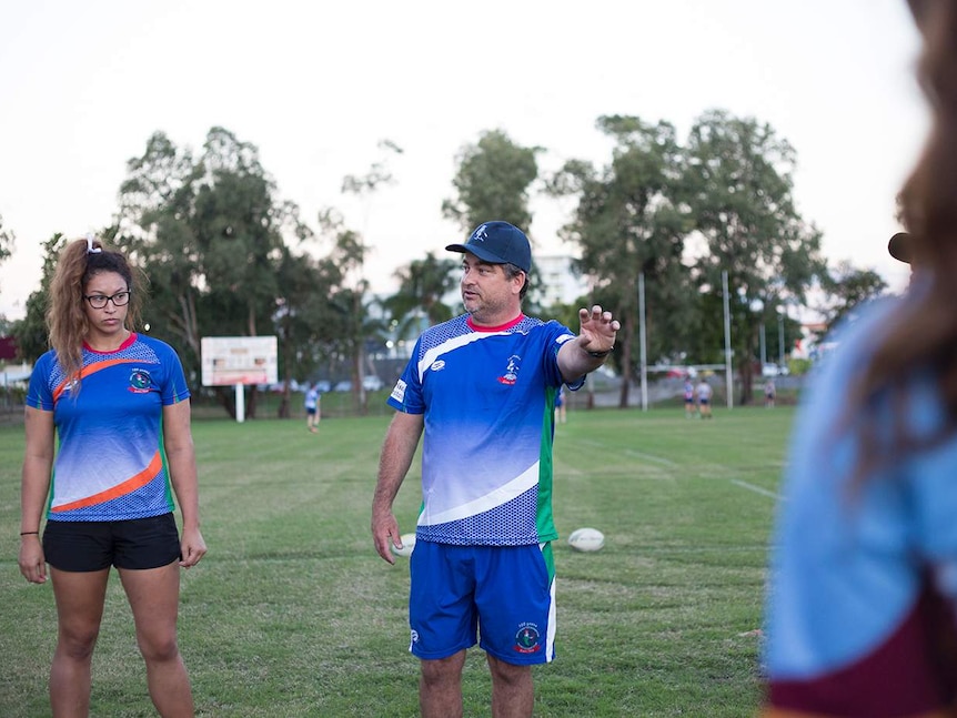 A coach stands between two players, talking and pointing on a rugby league field