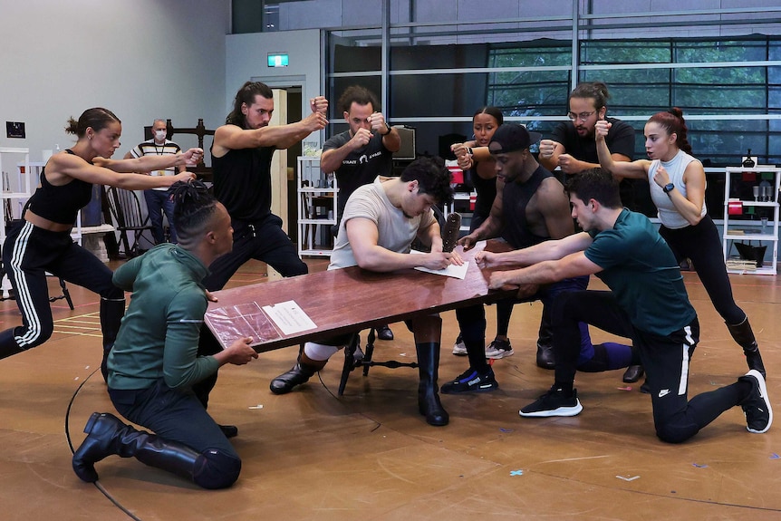 A group of people dance around a man writing on a plank of wood in a rehearsal studio.