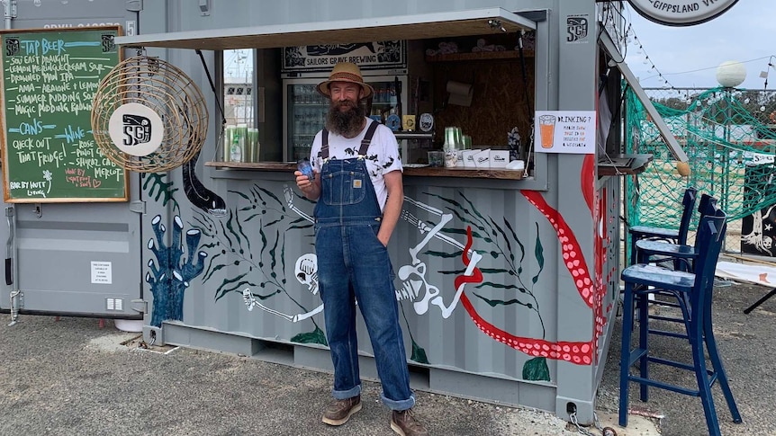 Man with hat and long beard holds a beer in front of shipping container stall.