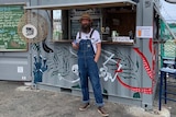 Man with hat and long beard holds a beer in front of shipping container stall.