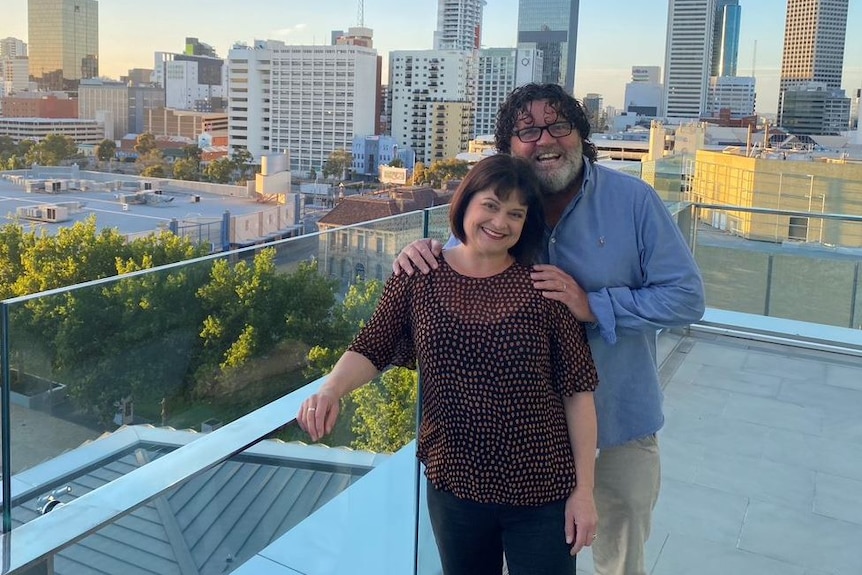 Nadia Mitsopoulos and Russell Woolf embrace and smile at the camera on a rooftop