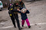 A man in a police uniform and combat helmet runs, holding the arms of a child guiding them as they run together.