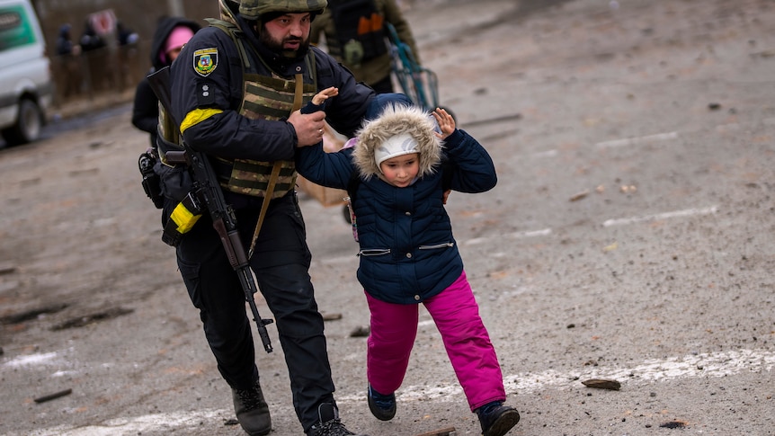 A man in a police uniform and combat helmet runs, holding the arms of a child guiding them as they run together.