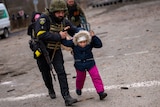 A man in a police uniform and combat helmet runs, holding the arms of a child guiding them as they run together.