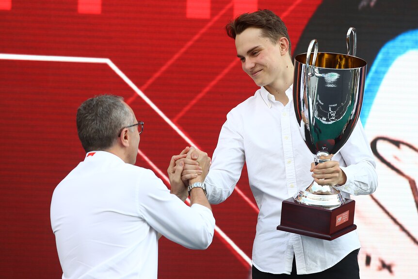 Australian driver Oscar Piastri shakes hands with the Formula One CEO as he holds a trophy after winning an F2 world title.