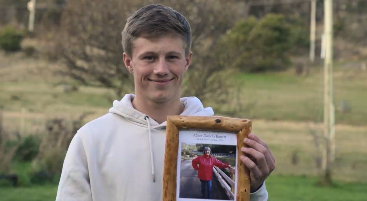 A young man in a white hoodie holding a photo frame of an older woman, smiling at the camera.