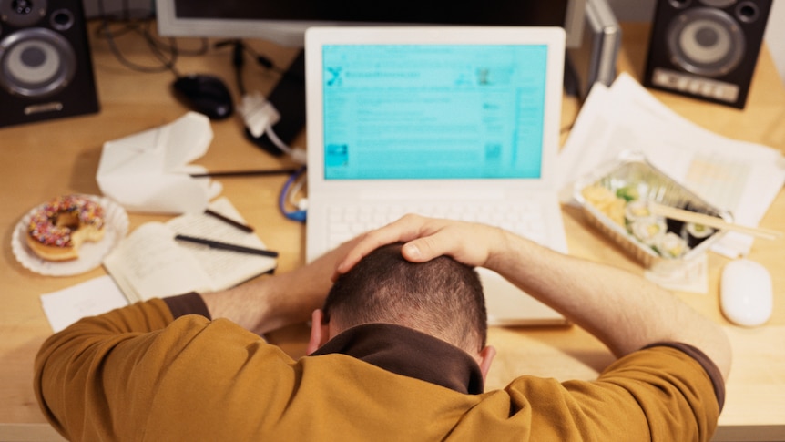 a man viewed from the back holding his head in his hands over his laptop surrounded by unhealthy food