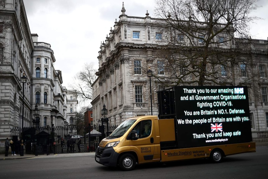 A gold van carrying a large screen displaying a message of support for the NHS is parked in front of neoclassical buildings.