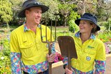 Two gardeners holding a shovel and pitchfork smile in a garden