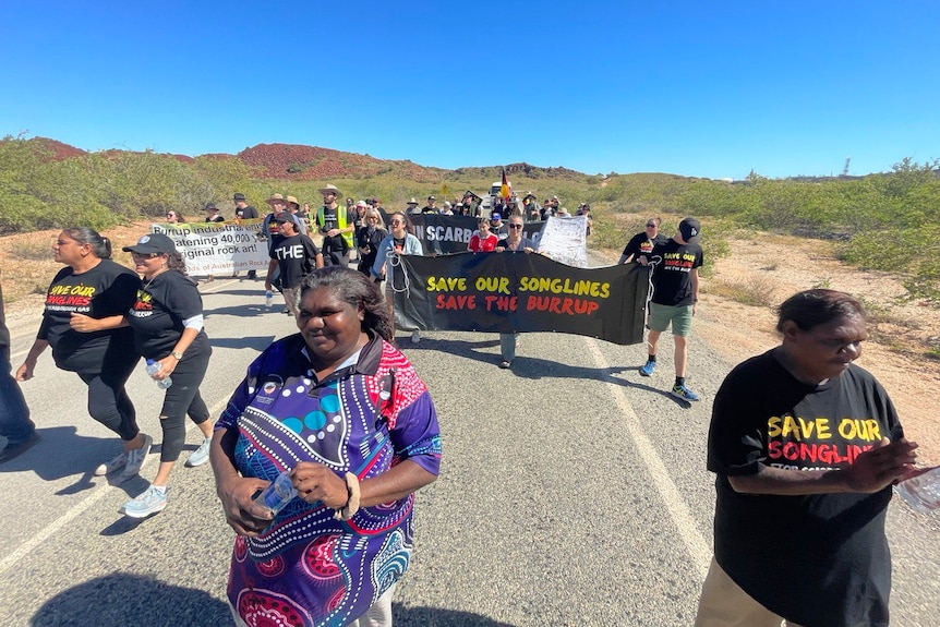 A group of people walk along a road in the outback holding signs