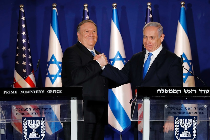 Standing in front of US and Israeli flags Mike Pompeo and Benjamin Netanyahu shake hands.