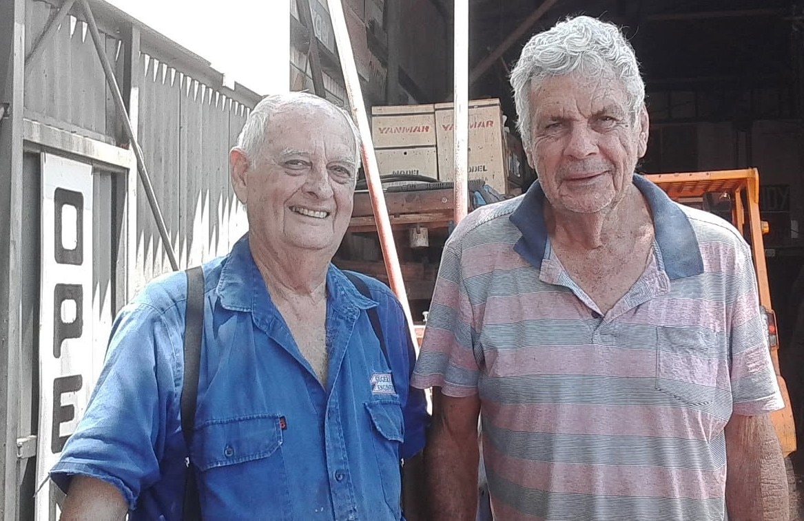 Two men in a shed smile at the camera