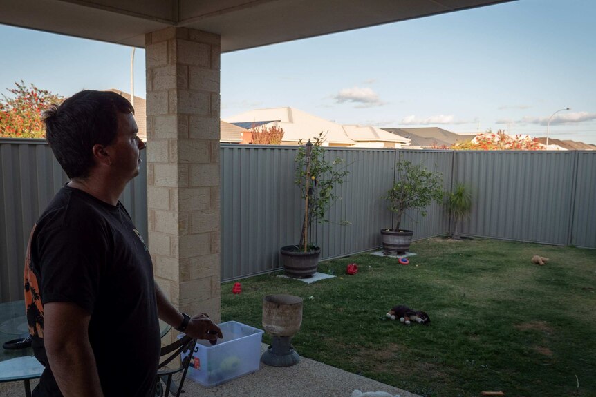 Man (Denis Grzetic) stands in porch in a backyard, looking over the fence and into the distance.