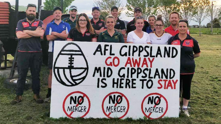 A group of country AFL footballers hold up a sign showing their opposition to the proposed merger of their league