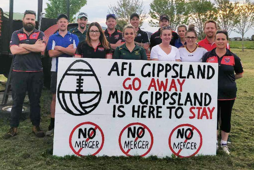 A group of country AFL footballers hold up a sign showing their opposition to the proposed merger of their league.