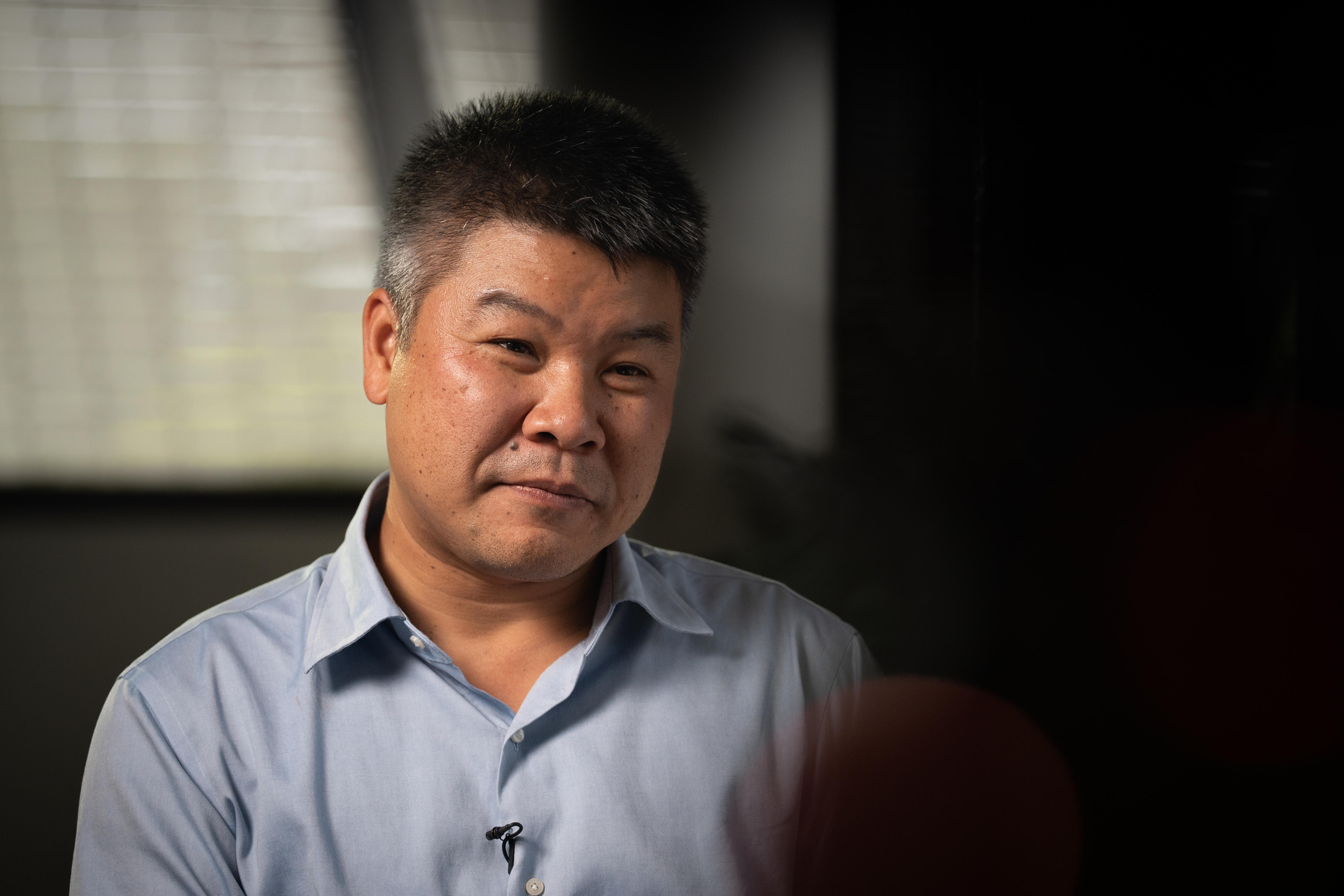 Um homem de Hong Kong olhando para uma pessoa à direita da câmera, cabelo preto/cinza, camisa de botão azul claro, expressão gentil e séria.