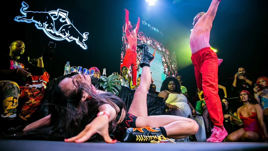 Vogue performer in red gloves and pants mid-move, while another kicks the air, during a vogue battle onstage at Sissy Ball 2019.