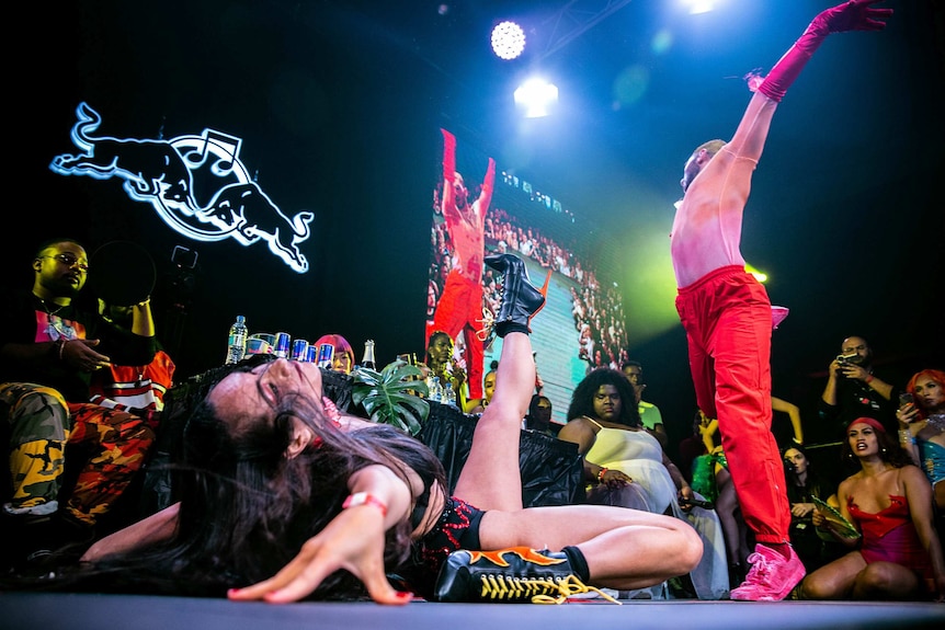 Vogue performer in red gloves and pants mid-move, while another kicks the air, during a vogue battle onstage at Sissy Ball 2019.