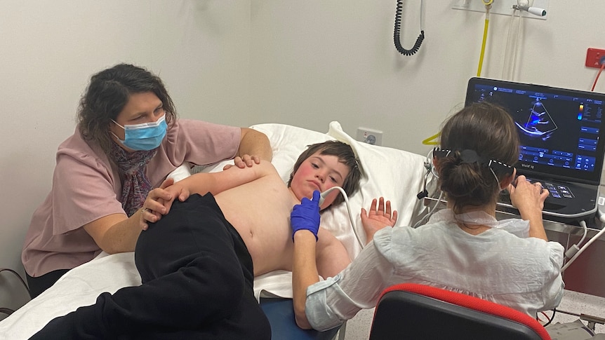 A shirtless boy lies on a hospital bed next to his mum, who is wearing a mask.