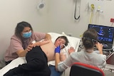 A shirtless boy lies on a hospital bed next to his mum, who is wearing a mask.