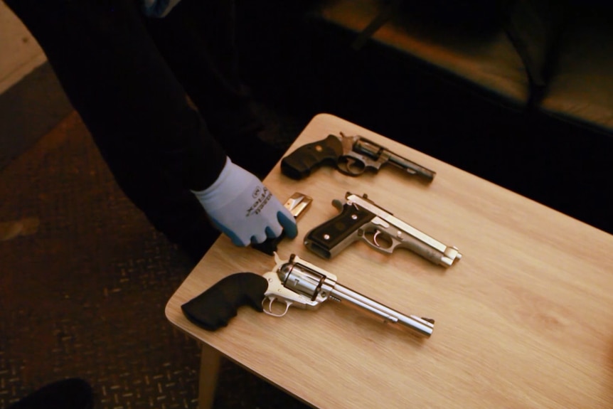 A small table with three guns on it, two revolvers and a handgun. A gloved hand is placing a clip of ammunition next to them.