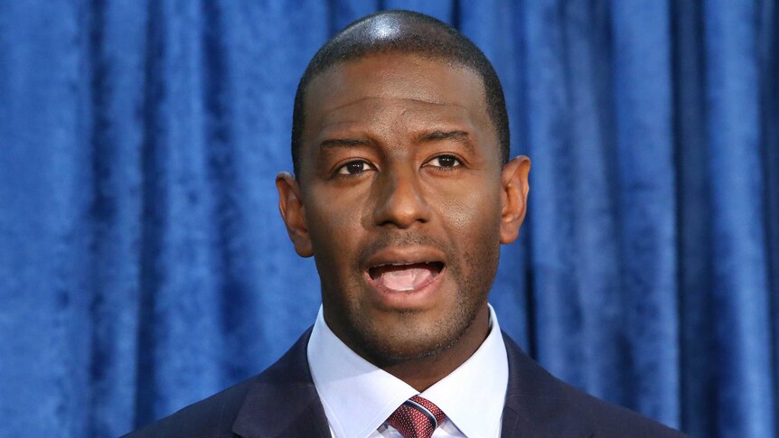 Andrew Gillum stands in a suit in front of a blue curtain, speaking into two microphones on stands in front of him