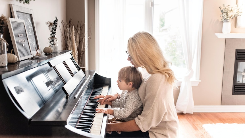 A blonde woman sitting at a piano with a young child on her lap who is pressing the keys