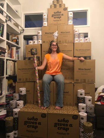 A woman with a makeshift staff and crown sitting atop a throne of toilet paper
