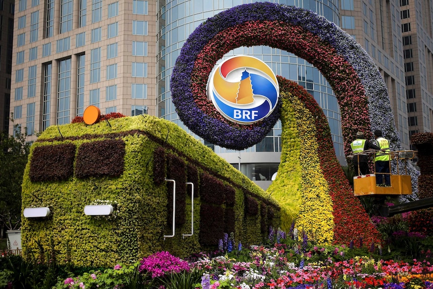 Workers stand on a platform installing flowers on a decoration in the shape of a train.