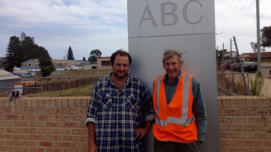 90 year old man in orange fluro vest stands outside next to younger man in flannelette shirt in front of ABC Esperance sign
