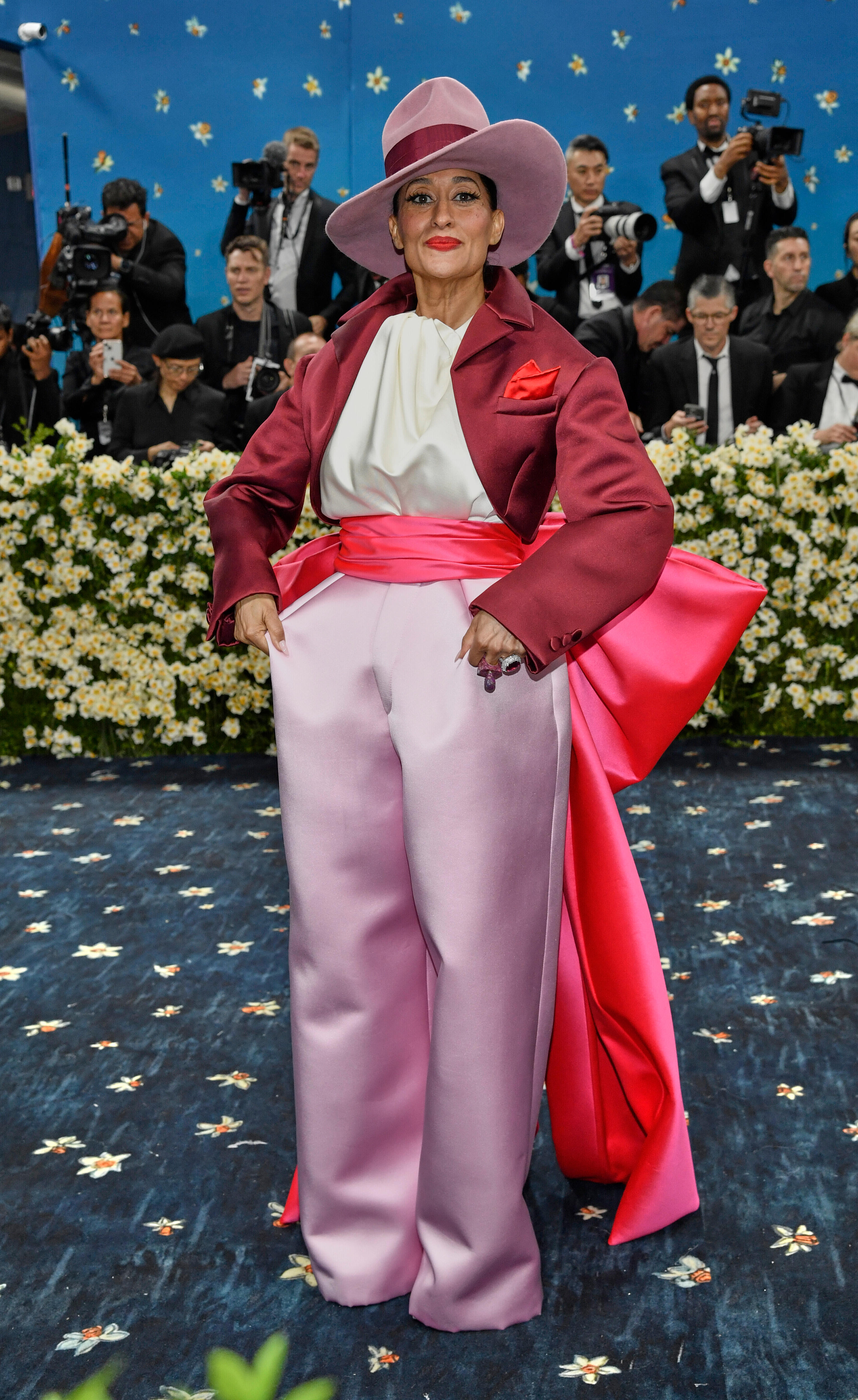 Tracee Ellis Ross wearing a big pink hat, silky pink trousers and a maroon jacket with a big bow on the back