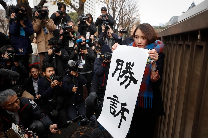 Shiori Ito holding up a sign with Japanese wording that translates to "winning the lawsuit". She is surrounded by cameras.