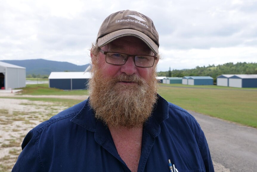 A man with a ginger beard in a dark grey cap and blue button up workman's shirt standing at an airfield.