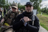 A man in black and cap gestures next to man in camouflage gear.