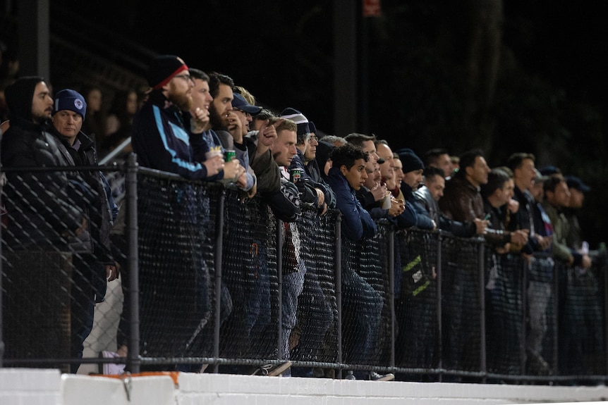 A group of people wearing winter clothes stands along a fenceline watching a sporting match