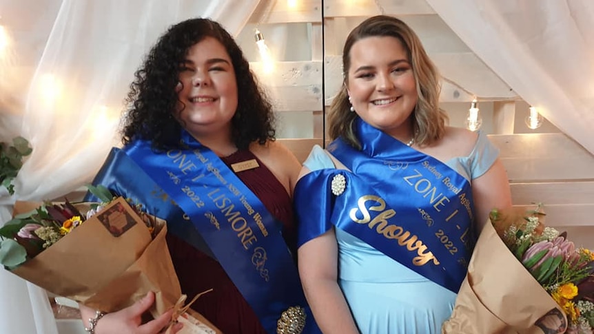 A young woman in a maroon dress stands next a young woman in a light blue dress. Both wear blue sashes and hold flowers.