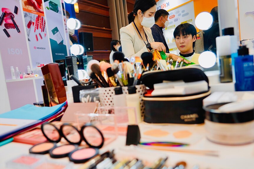 A Korean woman in a face mask talks to a man seated at a makeup counter, surrounded by cosmetics 
