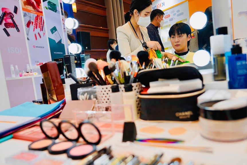 A Korean woman in a face mask talks to a man sitting at a makeup counter, surrounded by cosmetics 