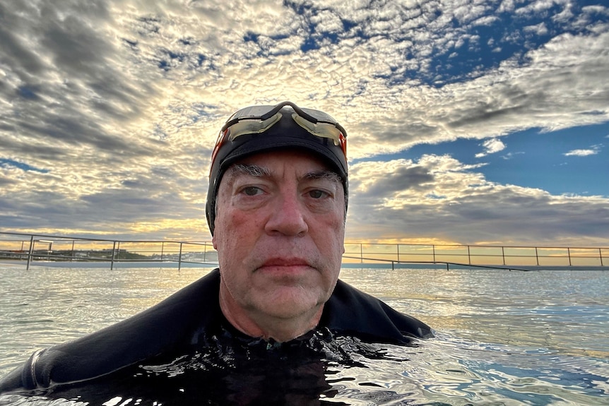 A man in a wetsuit takes a selfie with the sunrise in the background.