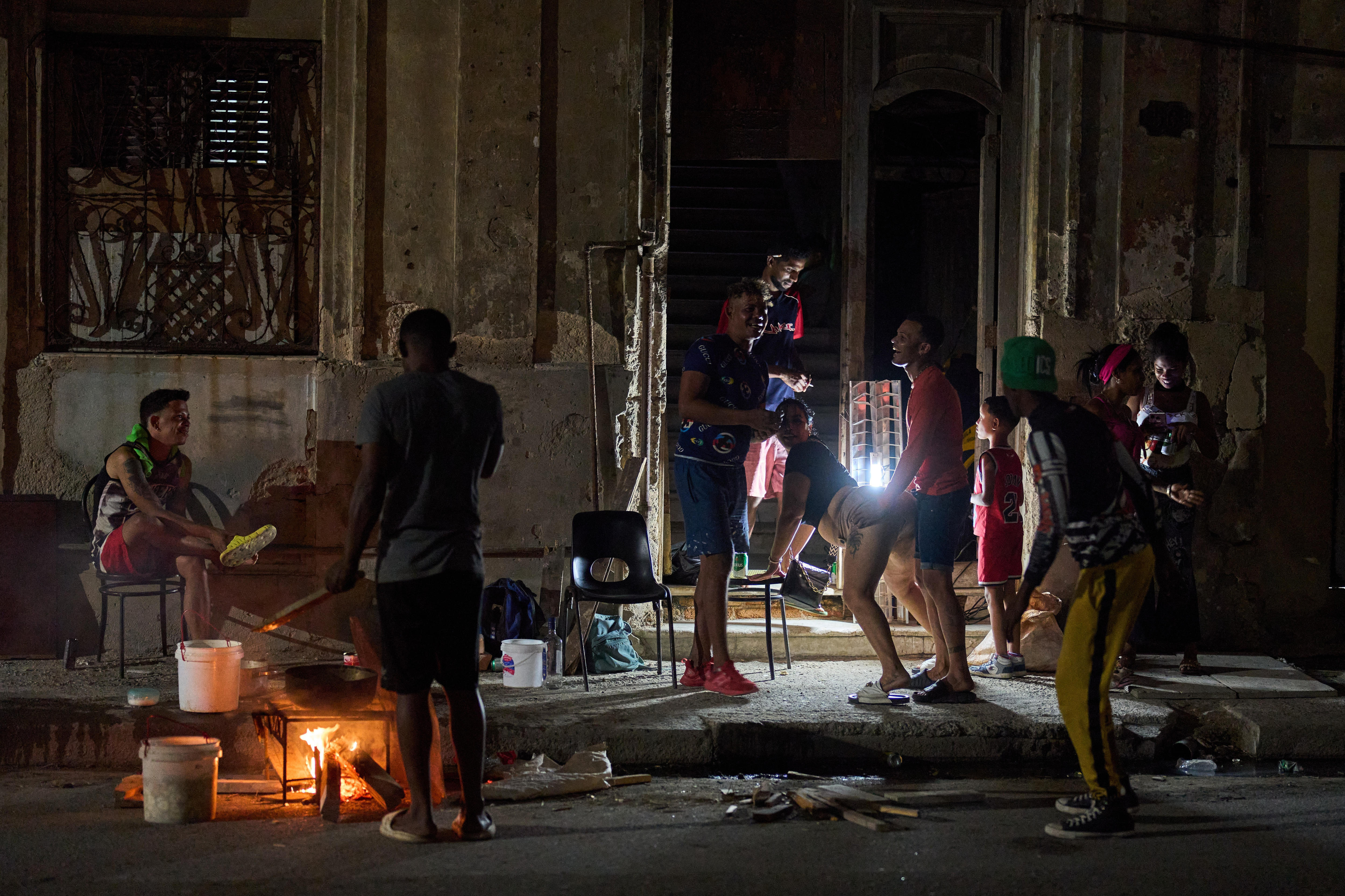 Hombres cubanos se sientan y se paran en una calle alrededor de una zona de cocina iluminada por un fuego y una lámpara halógena.
