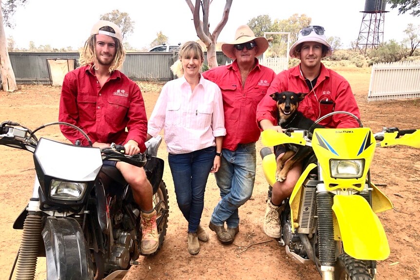 A woman in a pink shirt and a man in a red shirt stand on dusty ground, flanked on either side by two young men on motorbikes.