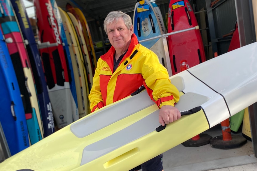 A man in a yellow and red surf life saving jacket holds a large paddle board