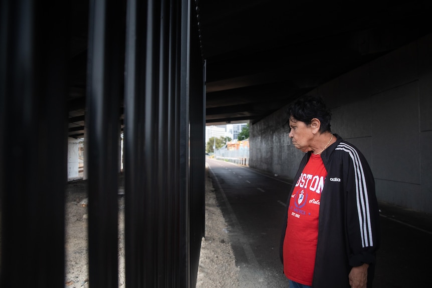 An Aboriginal woman stands side-on looking at a fence under an underpass.