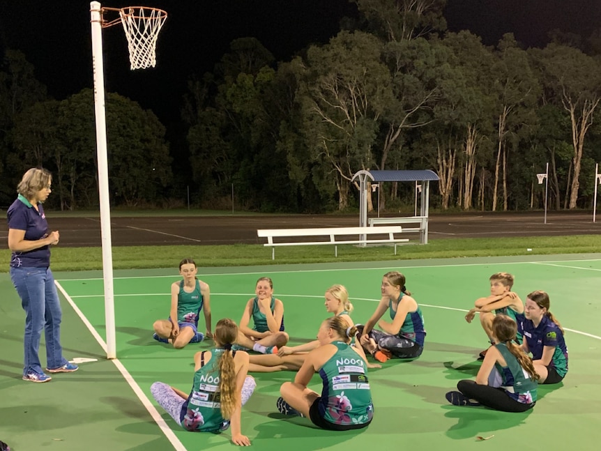 A team of young teenage netball players sit on a court, listening to the coach who is standing after an evening training session