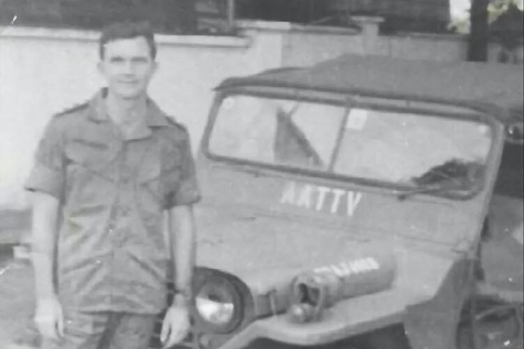 A black and white image of man in army fatigues in front of a jeep.