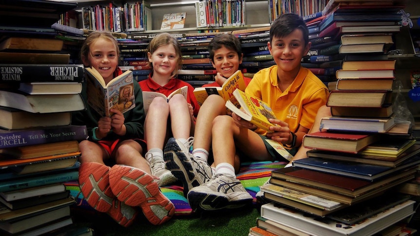 Four students sit on the floor reading books, while surrounded by piles of other books.