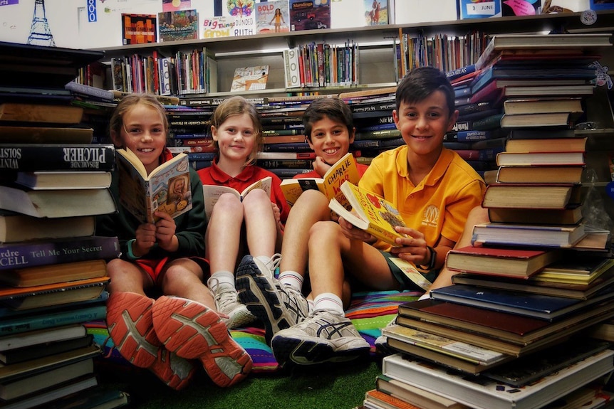 Four students sit on the floor reading books, while surrounded by piles of other books.