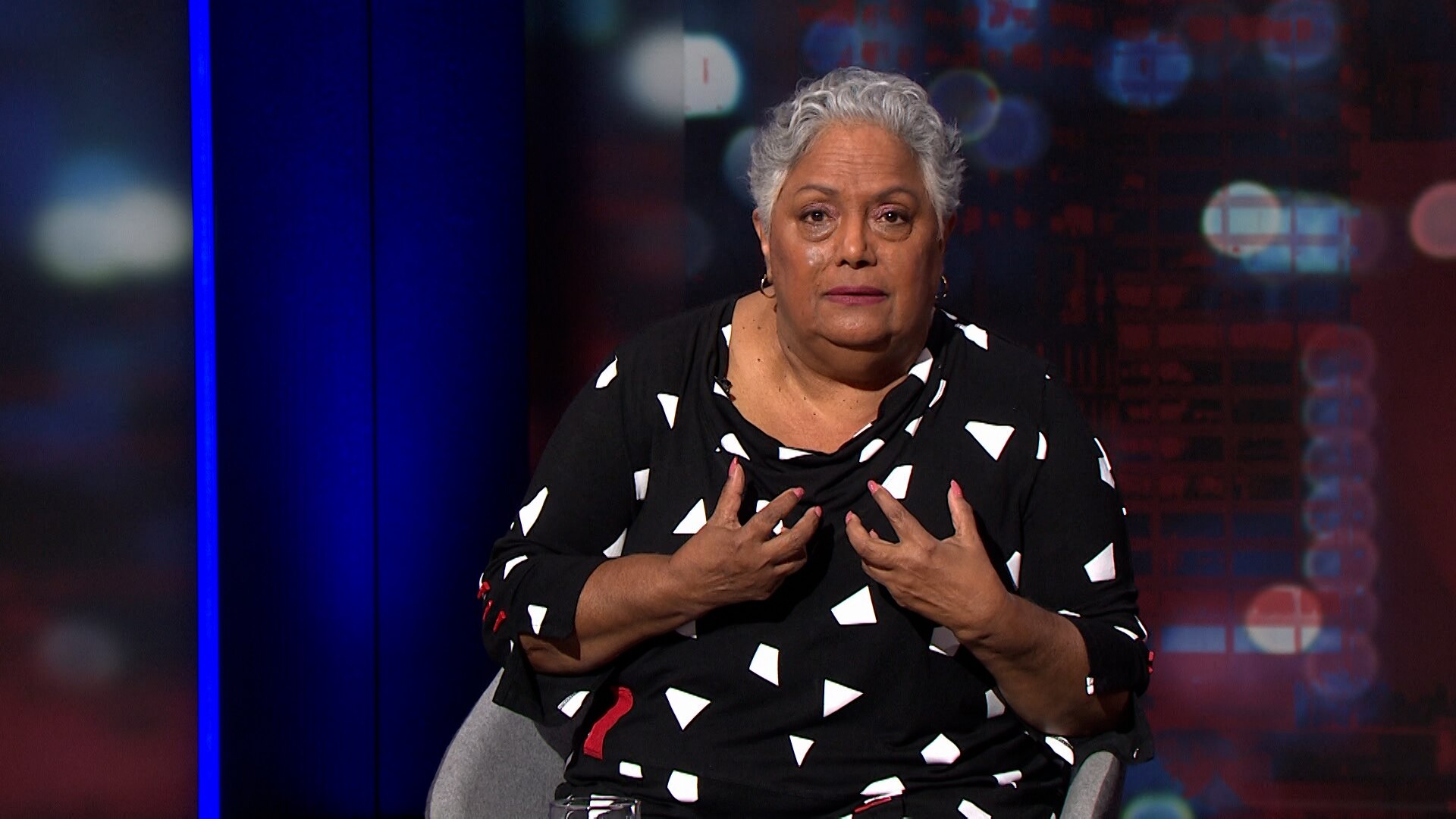 A woman in a black dress clutches her hands to her chest.